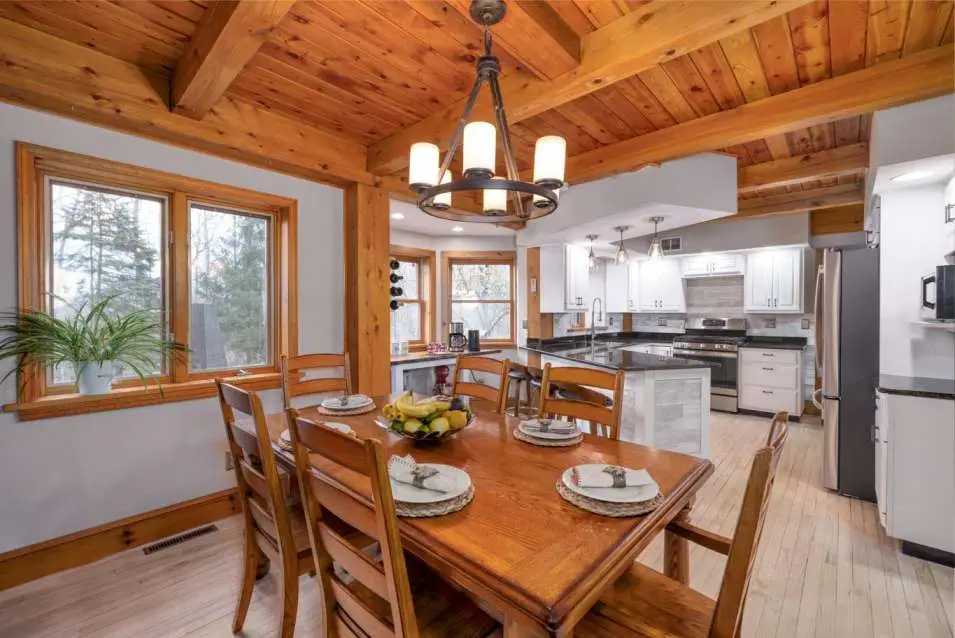 Interior dining area featuring a wooden table set with plates and a fruit bowl, surrounded by chairs, with large windows showcasing natural light and a rustic ceiling, highlighting the modern kitchen design in a real estate photography context.