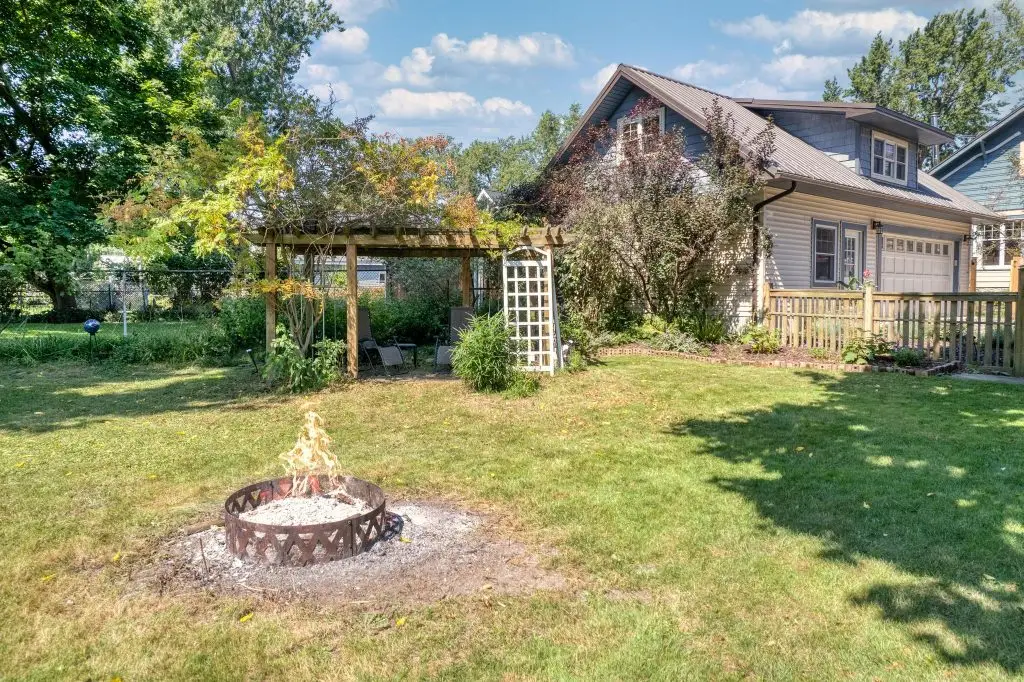 Backyard scene featuring a cozy fire pit surrounded by green grass, a wooden pergola with seating, and a charming house in West Seneca, NY, highlighting outdoor living potential for real estate listings.